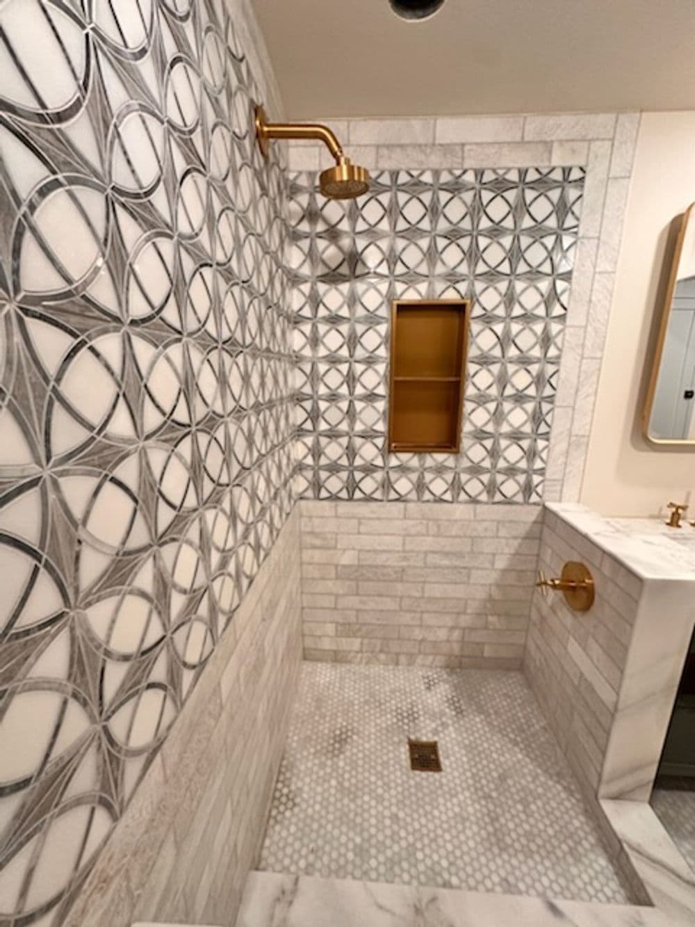 Modern bathroom with intricate marble tile wall, gold fixtures, and glass shower niche.